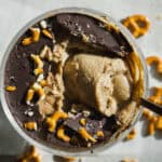 A glass cup filled with creamy dessert topped with a layer of dark chocolate and crushed pretzels, with a spoon scooping from the center; broken pretzel pieces are scattered around the cup.