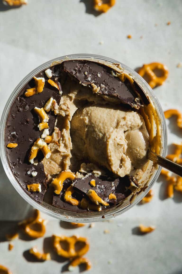 A close-up of a dessert cup with creamy sunflower butter mousse filling, a cracked chocolate layer on top, and scattered pieces of pretzel. A spoon is dug into the dessert, with more broken pretzel pieces around the cup on the surface.