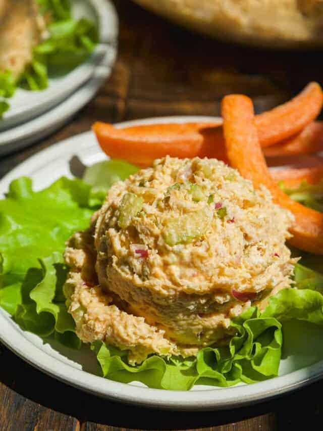 A scoop of chicken salad with chopped celery and red onion served on a bed of lettuce, accompanied by carrot sticks on a white plate.