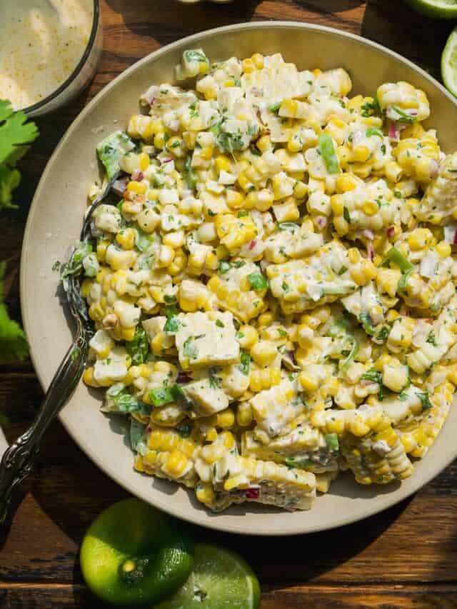 A bowl of creamy corn salad with chopped cilantro, red onions, and poblano peppers, served with lime wedges on the side. The salad appears fresh, colorful, and garnished with a light dressing.