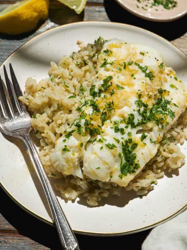 A plate of brown rice topped with a piece of white fish garnished with chopped parsley and lemon zest, with a fork on the side. A lemon wedge is in the background.