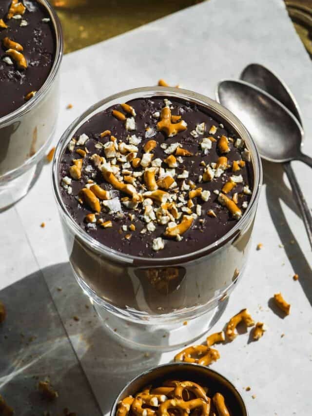 A glass filled with chocolate dessert topped with crushed pretzels, with two spoons and more pretzel pieces scattered on a white surface nearby.