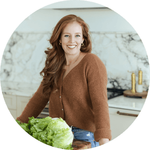 A woman with long red hair, wearing a brown sweater and jeans, smiles while standing in a modern kitchen beside a counter with fresh lettuce and hibachi steak bowls. Marble backsplash and gold kitchen accents are visible in the background.