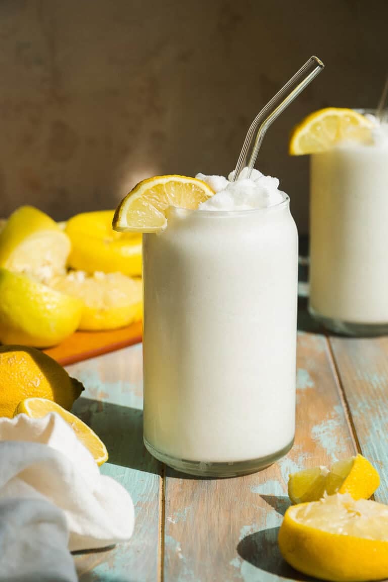 A frosty glass of white frozen lemonade with a glass straw and lemon slice garnish sits on a wooden table. Whole and sliced lemons and another glass of the drink are in the background. Sunlight shines on the scene.
