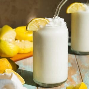 A frosty glass of lemon slush, garnished with a lemon slice and a glass straw, sits on a wooden table with fresh lemons and another drink in the background. Sunlight highlights the creamy, refreshing texture.
