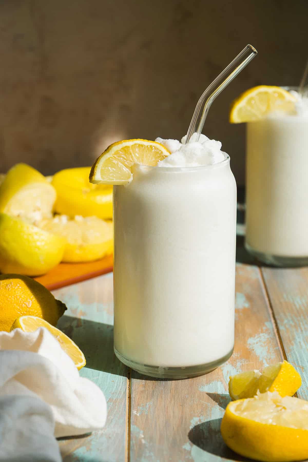 A frosty glass of lemon slush, garnished with a lemon slice and a glass straw, sits on a wooden table with fresh lemons and another drink in the background. Sunlight highlights the creamy, refreshing texture.
