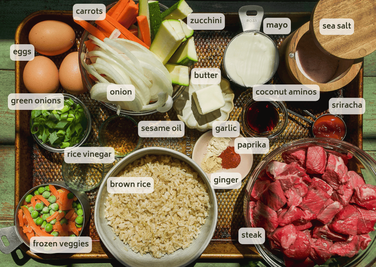 A tray with labeled ingredients for hibachi steak bowls: carrots, zucchini, eggs, mayo, sea salt, green onions, onion, butter, coconut aminos, sriracha, sesame oil, garlic, paprika, ginger, rice vinegar, brown rice, steak and frozen veggies.