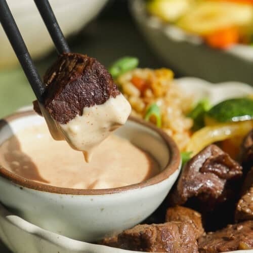 A piece of cooked steak held by chopsticks is being dipped into creamy sauce, with rice, vegetables, and more hibachi steak bowls arranged nearby.