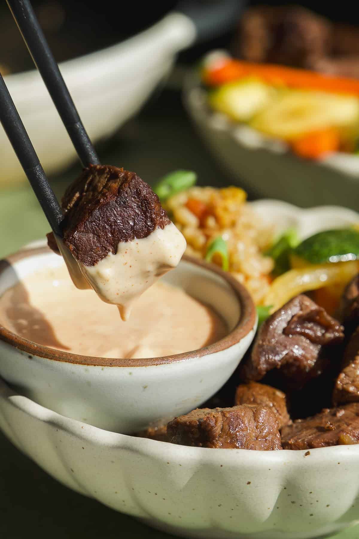 Chopsticks hold a piece of grilled beef dipped in creamy sauce above a small bowl, with vegetables and more beef pieces in the background.