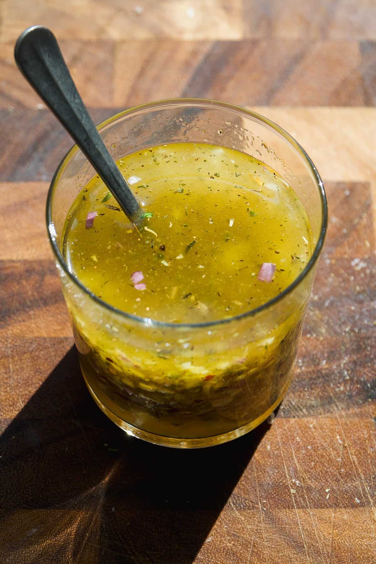 A glass filled with yellow salad dressing containing herbs and small pieces of red onion, with a metal spoon inside, placed on a wooden surface.