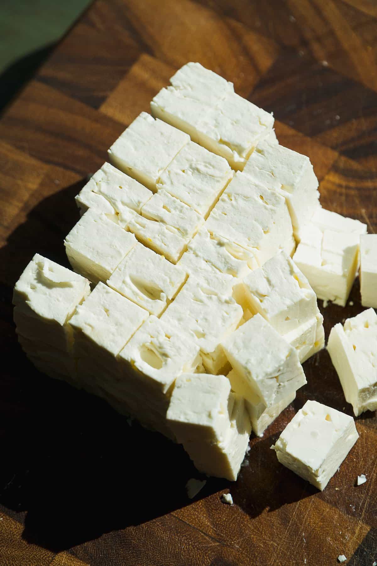 A block of white cheese cut into small cubes is arranged on a wooden cutting board, with sunlight casting shadows on the surface.