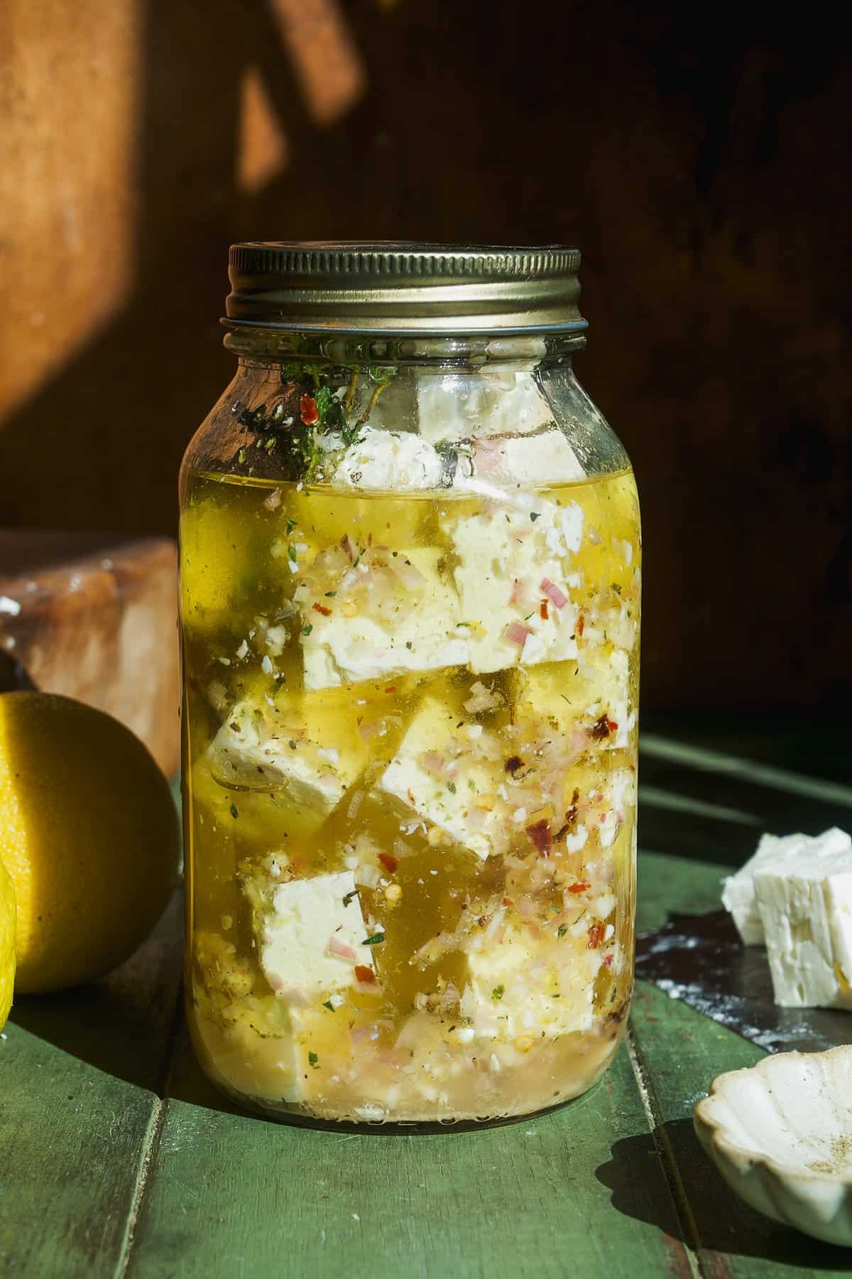 A glass jar filled with cubed feta cheese marinated in olive oil and herbs, placed on a green wooden surface with sunlight casting shadows; some cheese and a lemon are next to the jar.