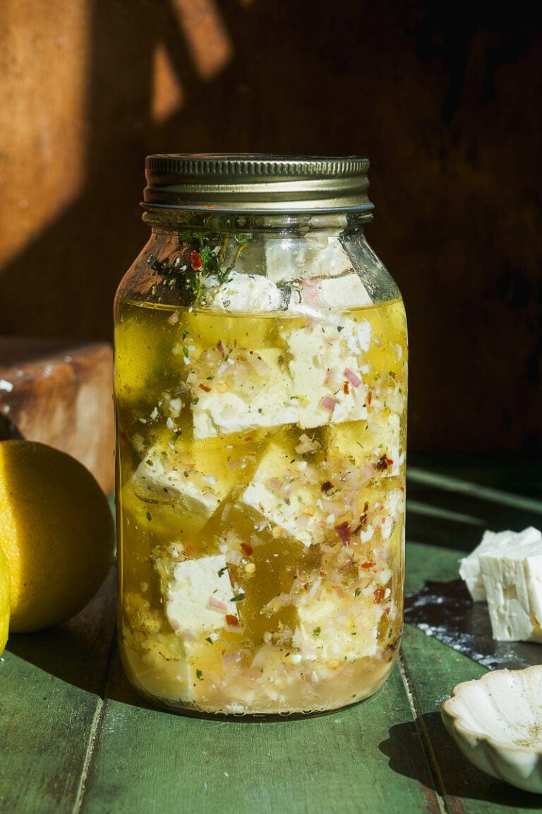 A glass jar filled with cubes of feta cheese marinating in olive oil with herbs and spices, sitting on a green wooden surface in warm sunlight.