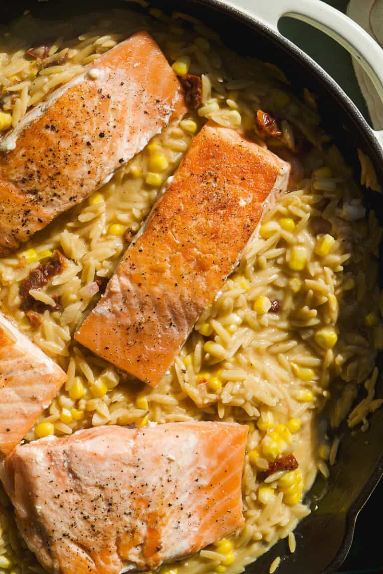 Three seared salmon fillets rest on creamy corn orzo pasta, mixed with sun-dried tomatoes in a skillet. The dish is garnished with black pepper and appears warm and freshly cooked.