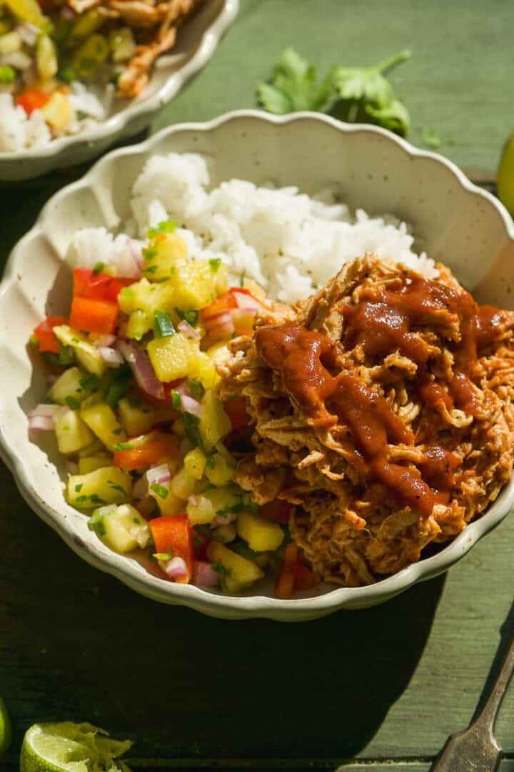 A bowl filled with white rice, colorful pineapple salsa with diced vegetables, and shredded bbq chicken topped with red sauce, set on a green surface.
