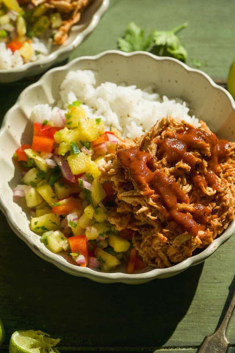 A bowl filled with white rice, colorful pineapple salsa with diced vegetables, and shredded bbq chicken topped with red sauce, set on a green surface.