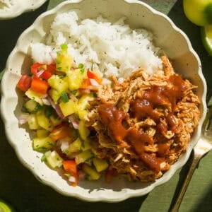 A bowl filled with white rice, shredded BBQ chicken topped with sauce, and colorful salsa of pineapple, tomato, and cucumber. Lime wedges and fresh herbs are nearby on a green wooden surface.