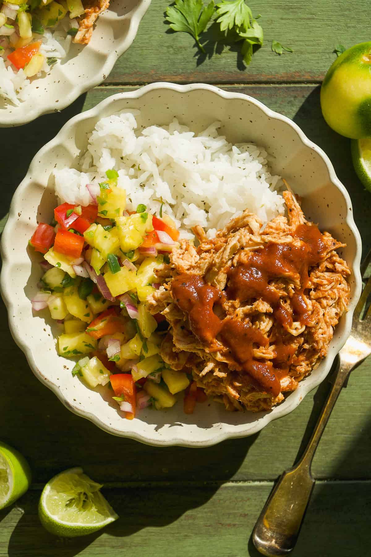A bowl filled with white rice, shredded BBQ chicken topped with sauce, and colorful salsa of pineapple, tomato, and cucumber. Lime wedges and fresh herbs are nearby on a green wooden surface.