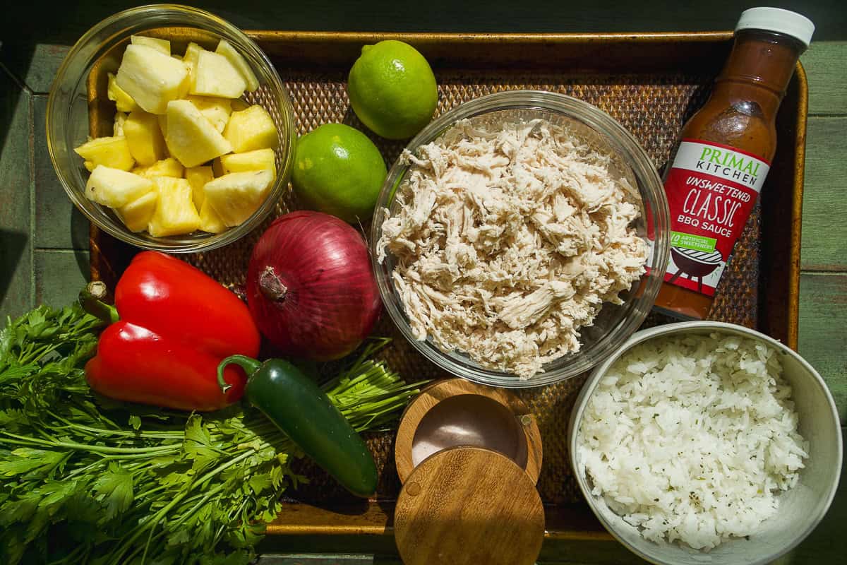 A tray with fresh ingredients for bbq chicken bowls: pineapple chunks, two limes, shredded chicken, red onion, red pepper, jalapeño, cilantro, a bowl of white rice, a wooden salt cellar, and classic BBQ sauce.