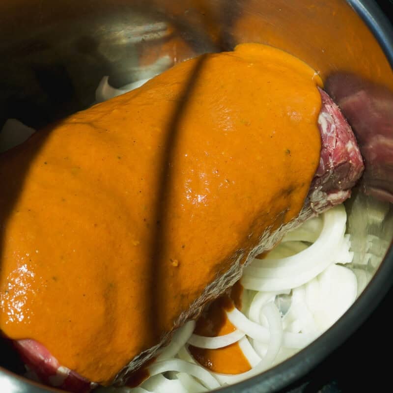 A raw beef roast sits on a bed of sliced onions in a metal pot, covered with a layer of thick orange sauce, ready for cooking—reminiscent of the savory flavors found in a bbq pulled pork bowl. Sunlight casts shadows across the ingredients.