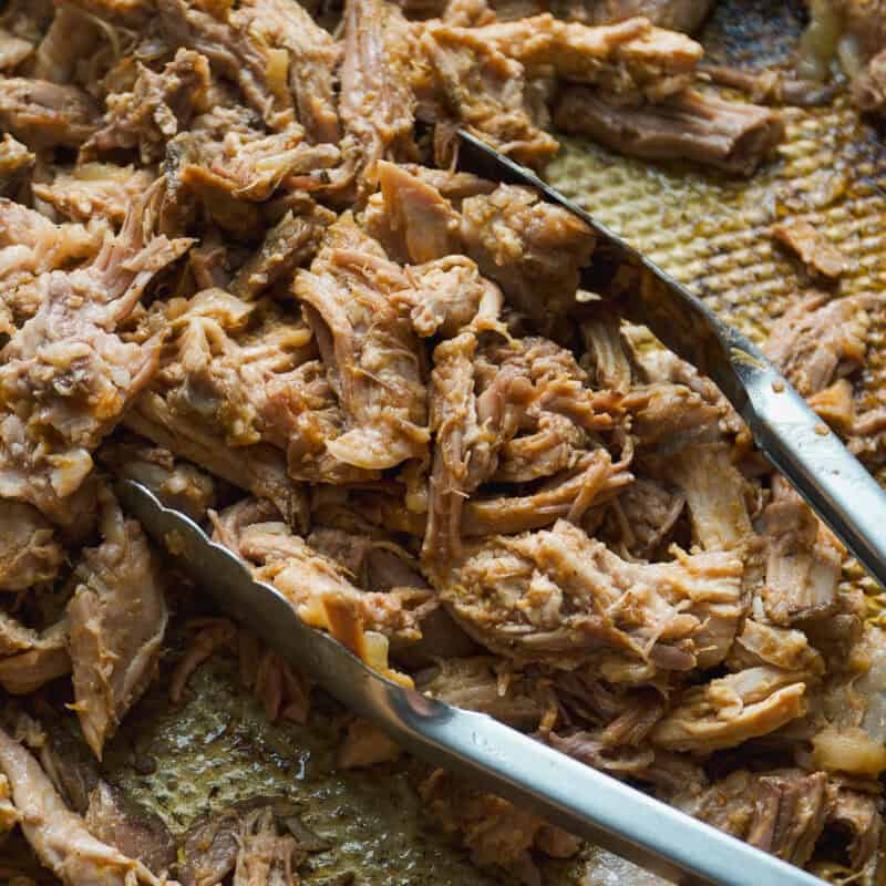 Shredded cooked meat on a tray with a pair of metal tongs resting on top, perfect for creating your own bbq pulled pork bowl.