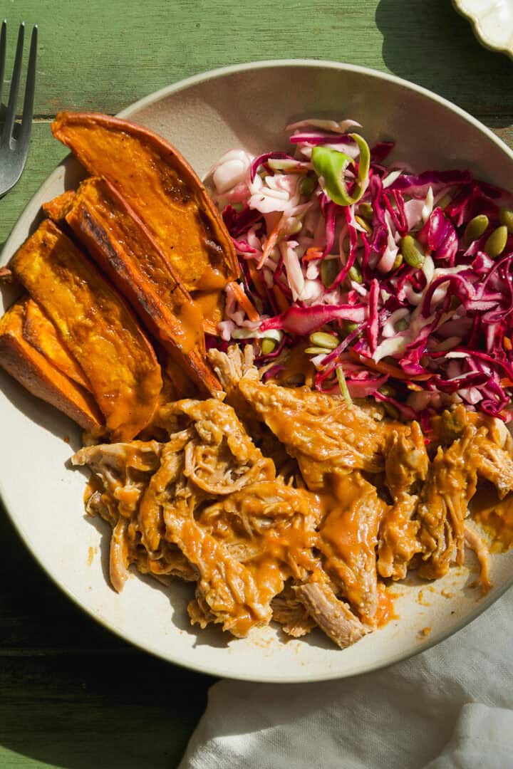A plate with bbq pulled pork bowl in sauce, roasted sweet potato wedges, and a colorful red cabbage slaw with seeds, served on a green table.