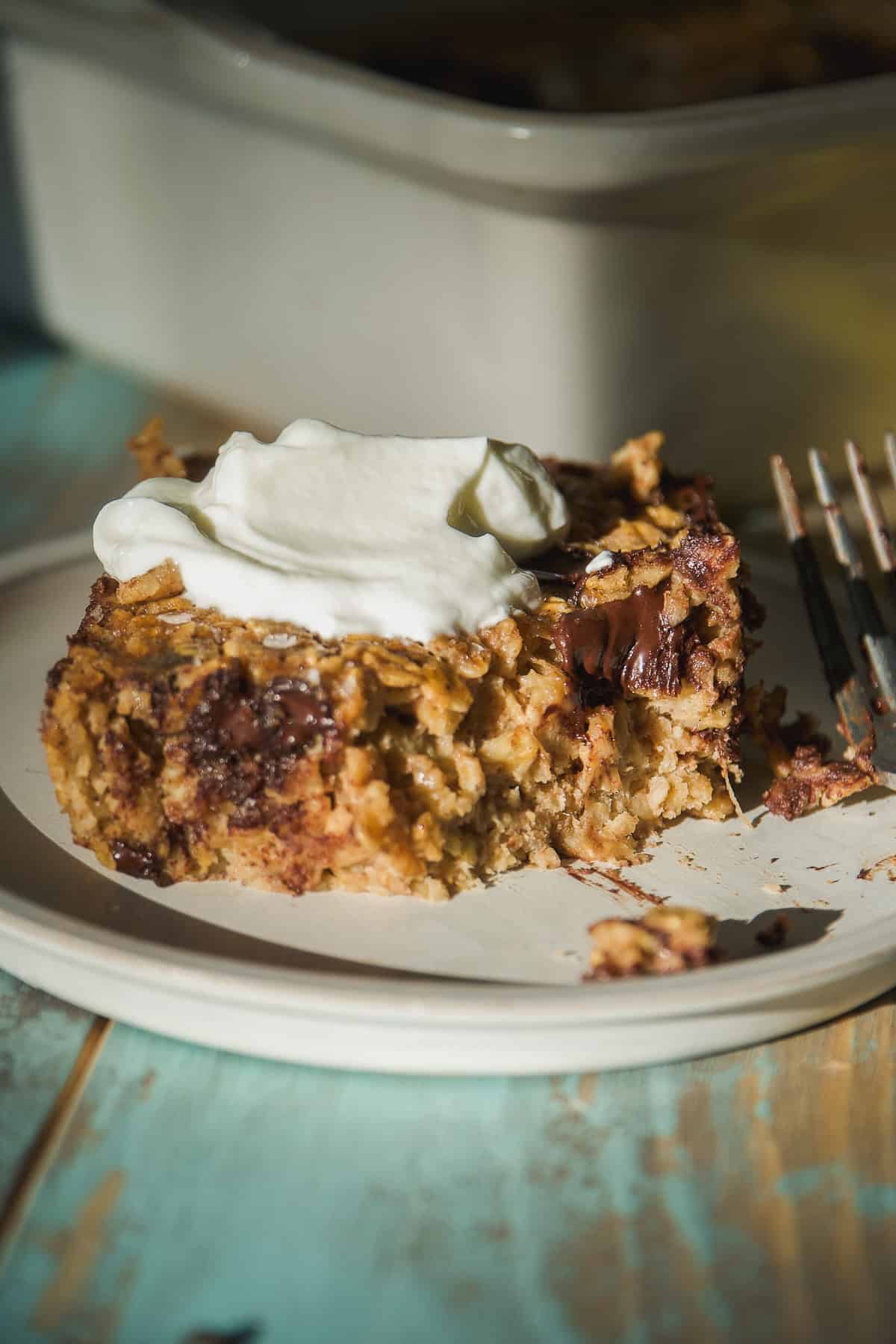 A close-up of a partially eaten banana bread baked oats bar with melted chocolate chunks, topped with a dollop of whipped cream and served on a white plate with a fork nearby.