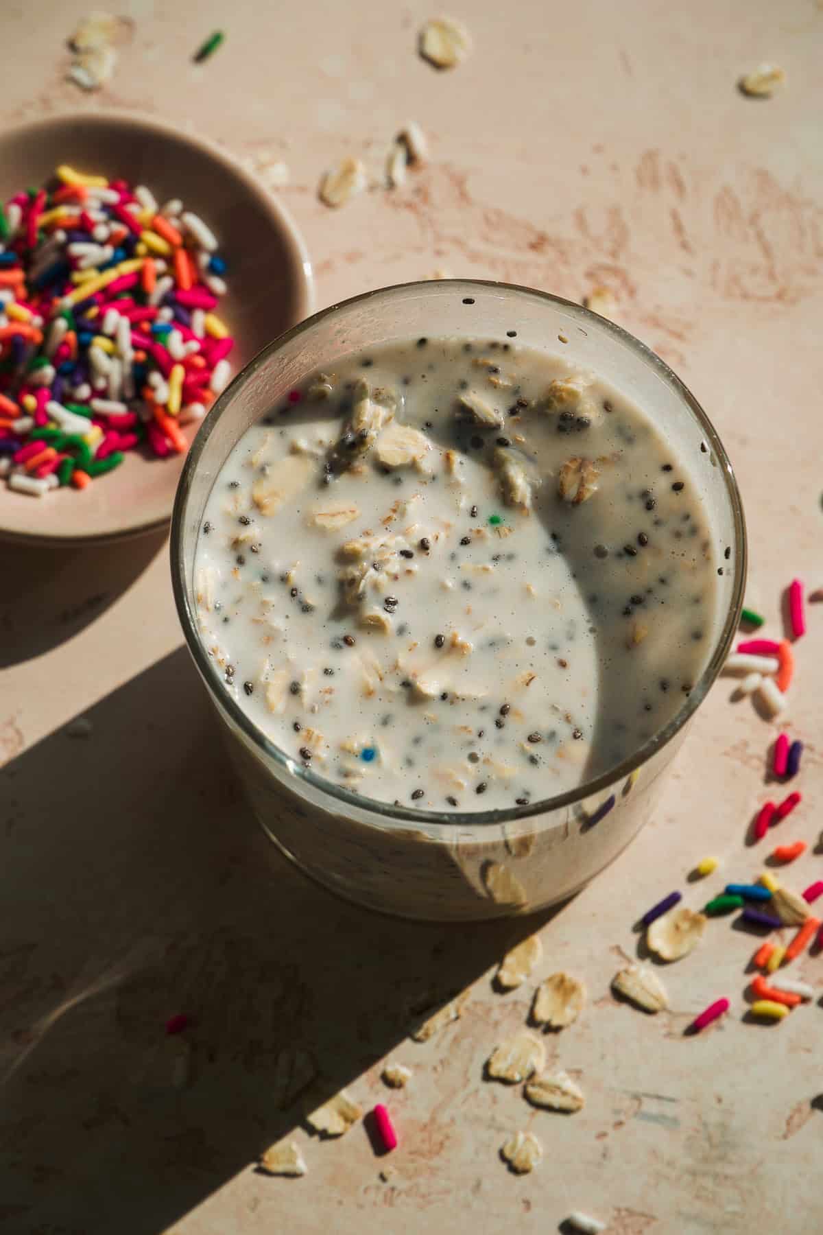 A glass of overnight oats with chia seeds and colorful sprinkles sits on a light surface, next to a small plate filled with more rainbow sprinkles and scattered oats and sprinkles around.