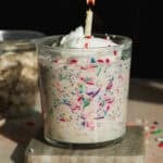 A glass jar filled with overnight oats mixed with colorful sprinkles, topped with whipped cream and a lit birthday candle, sits on a stone coaster with sprinkles scattered around.