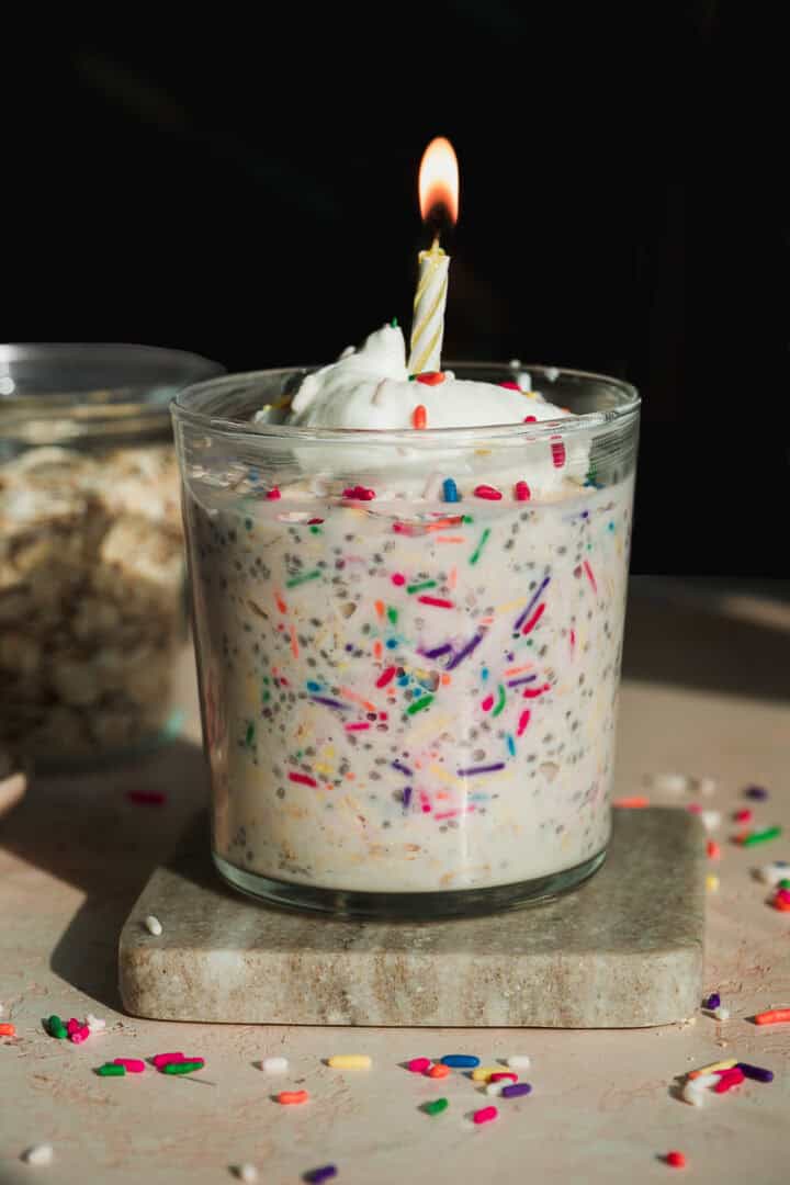 A glass filled with creamy oats mixed with colorful sprinkles, topped with whipped cream and a lit birthday candle. The glass sits on a stone coaster, with extra sprinkles scattered around.
