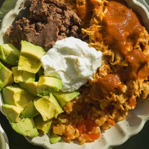 A bowl with shredded chicken in red sauce, refried beans, avocado chunks, Mexican rice, and a dollop of sour cream—this vibrant Mexican rice bowl is perfectly arranged side by side. A fork and a halved avocado are nearby.