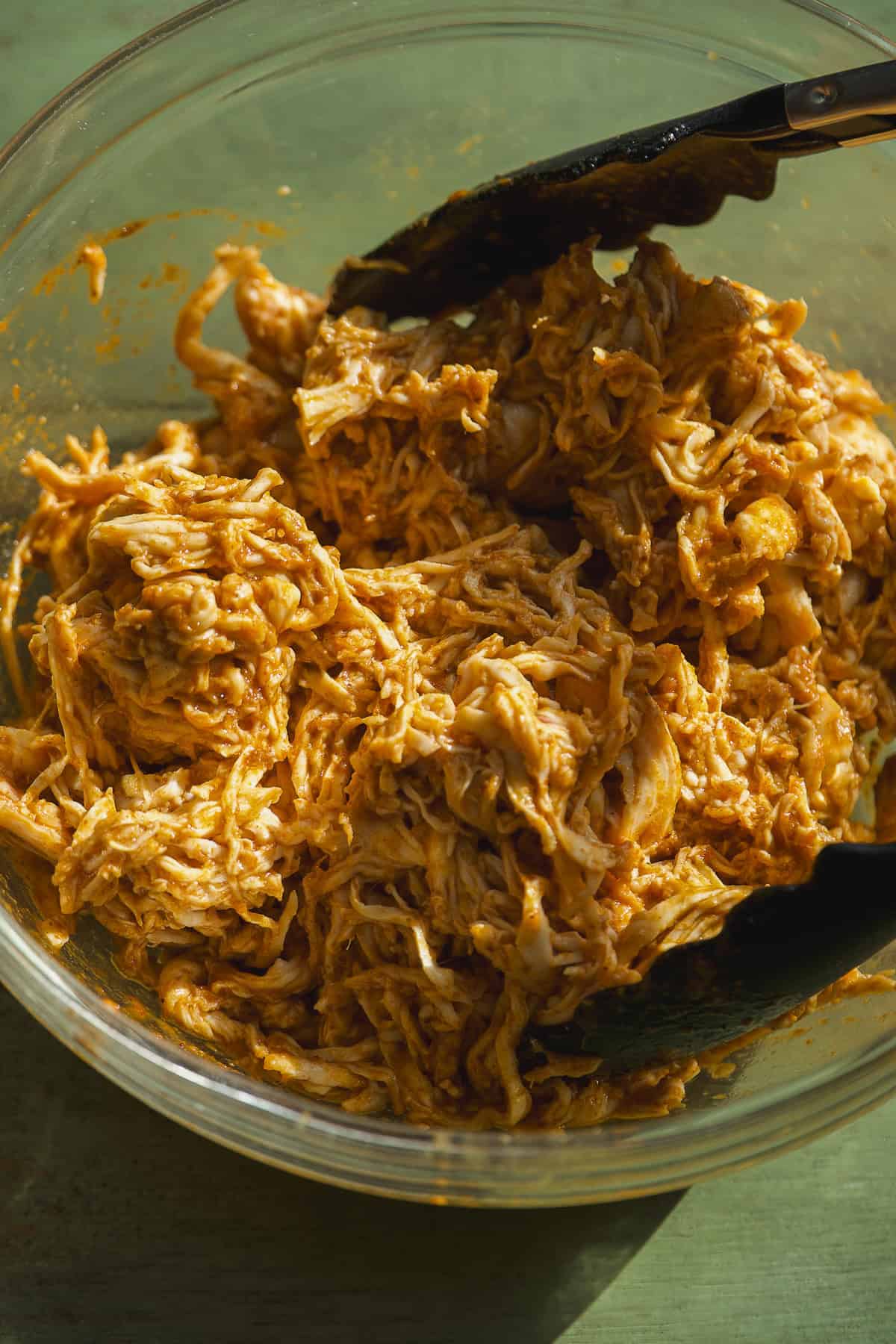 A glass bowl filled with shredded chicken coated in a creamy, orange-colored sauce, perfect for Mexican rice bowls, with a pair of black tongs resting in the mixture. The bowl sits on a green surface.