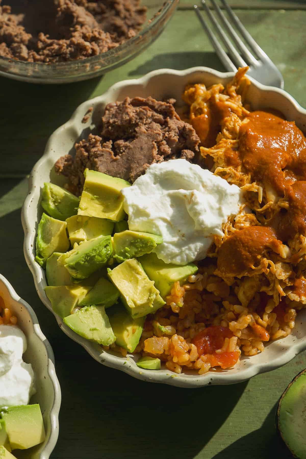 A delicious Mexican rice bowl featuring diced avocado, refried beans, shredded chicken in red sauce, tomato-infused Mexican rice, and a dollop of sour cream. A fork and another bowl are set nearby for easy dining.