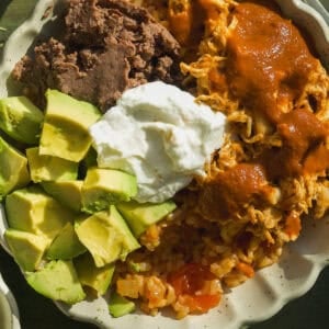 A delicious Mexican rice bowl featuring diced avocado, refried beans, shredded chicken with red sauce, Mexican rice, and a dollop of sour cream, all served in a scalloped dish on a green surface.