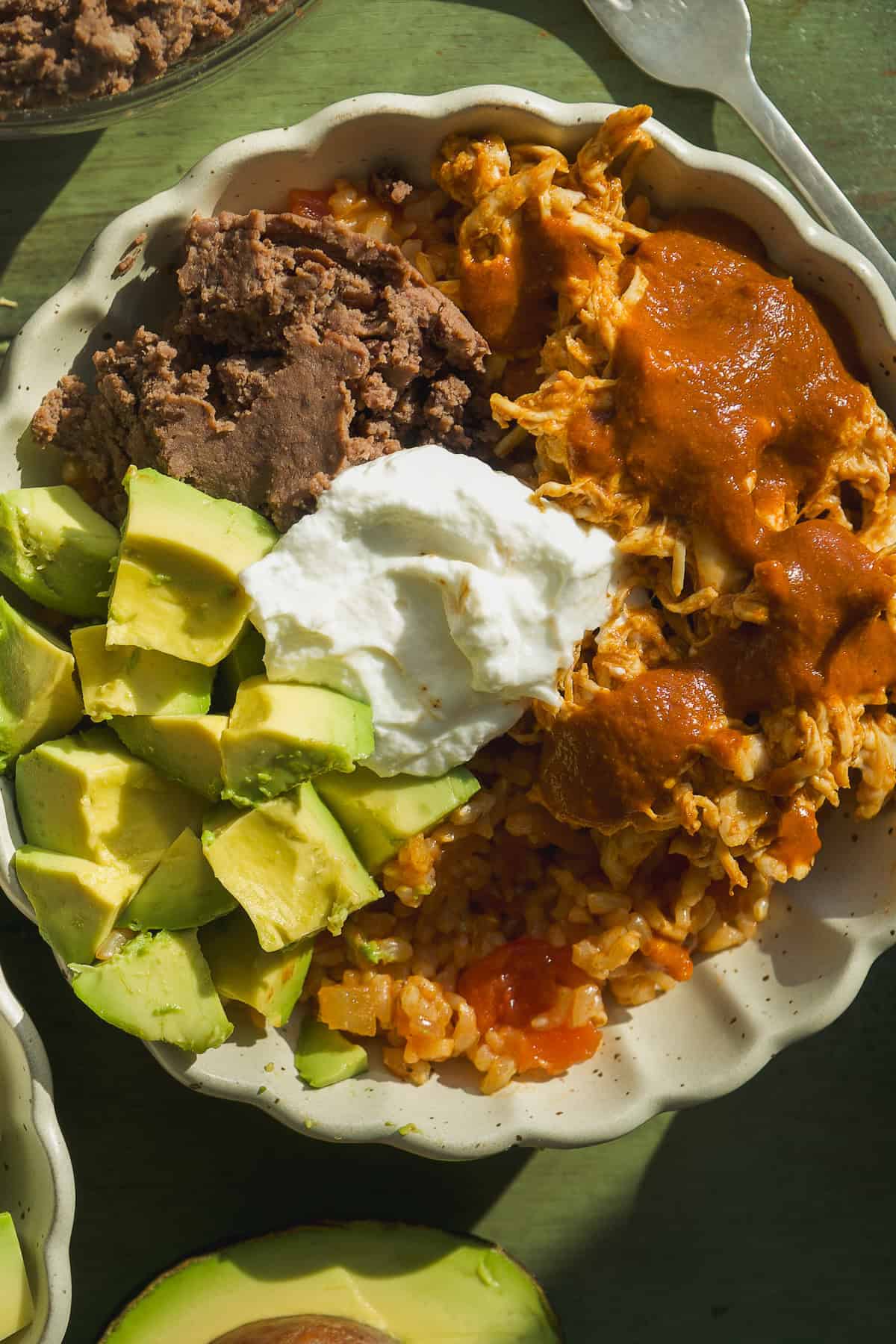 A delicious Mexican rice bowl featuring diced avocado, refried beans, shredded chicken with red sauce, Mexican rice, and a dollop of sour cream, all served in a scalloped dish on a green surface.