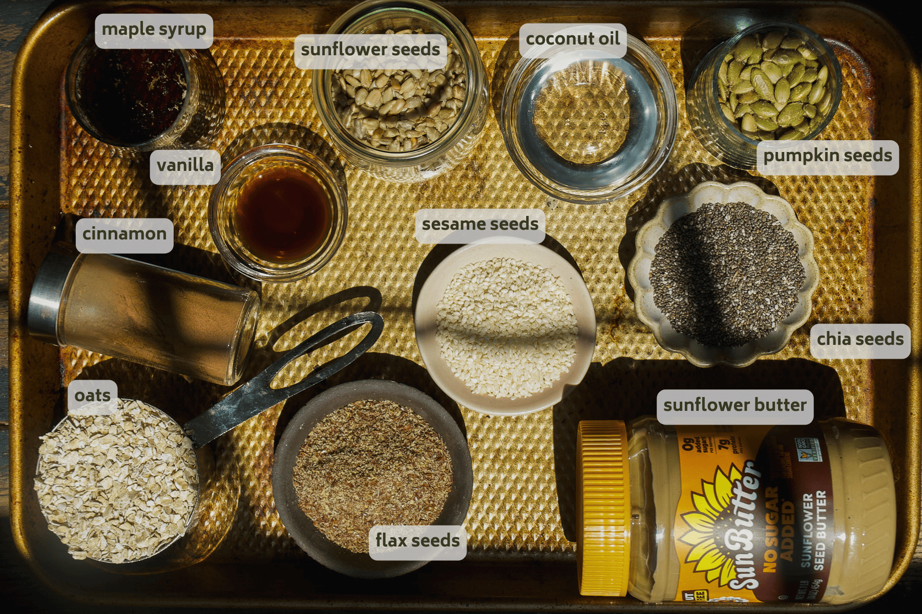 A baking tray holds small bowls and containers with labeled ingredients: maple syrup, sunflower seeds, coconut oil, pumpkin seeds, vanilla, cinnamon, oats, flax seeds, sesame seeds, sunflower butter, and chia seeds.