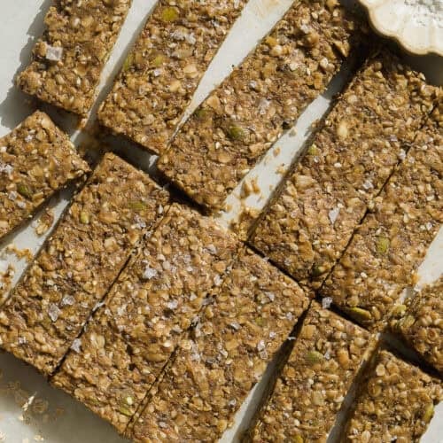 Homemade healthy breakfast bars arranged on parchment paper, sprinkled with sea salt. A small dish of flaky salt and a jar of rolled oats are nearby. The bars are packed with oats, seeds, and nuts.