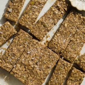 Homemade granola bars cut into rectangular pieces on parchment paper. The bars contain oats, seeds, and nuts, and are sprinkled with sea salt. A small dish of salt and a jar of oats are nearby.
