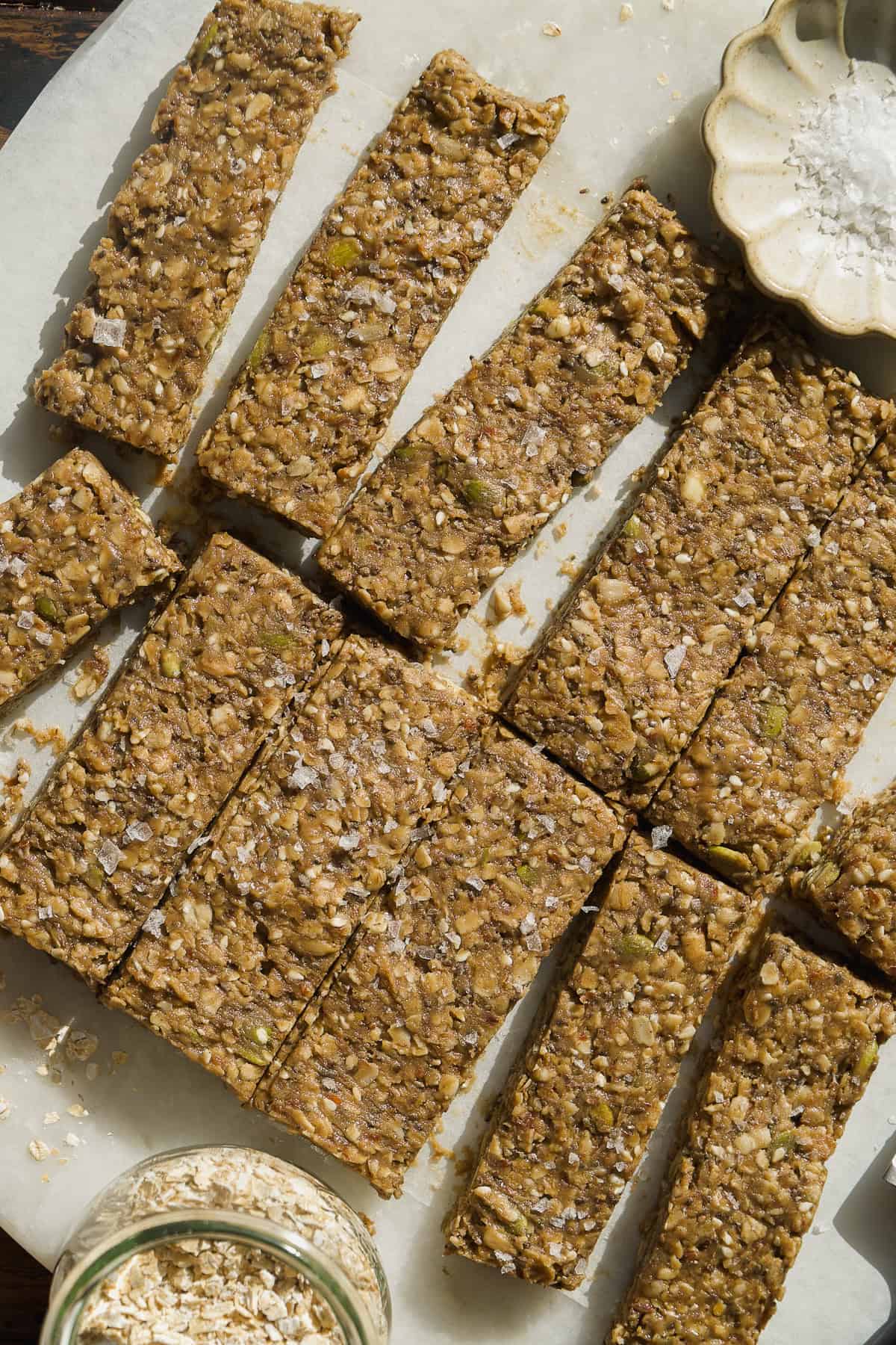 Homemade granola bars cut into rectangular pieces on parchment paper. The bars contain oats, seeds, and nuts, and are sprinkled with sea salt. A small dish of salt and a jar of oats are nearby.