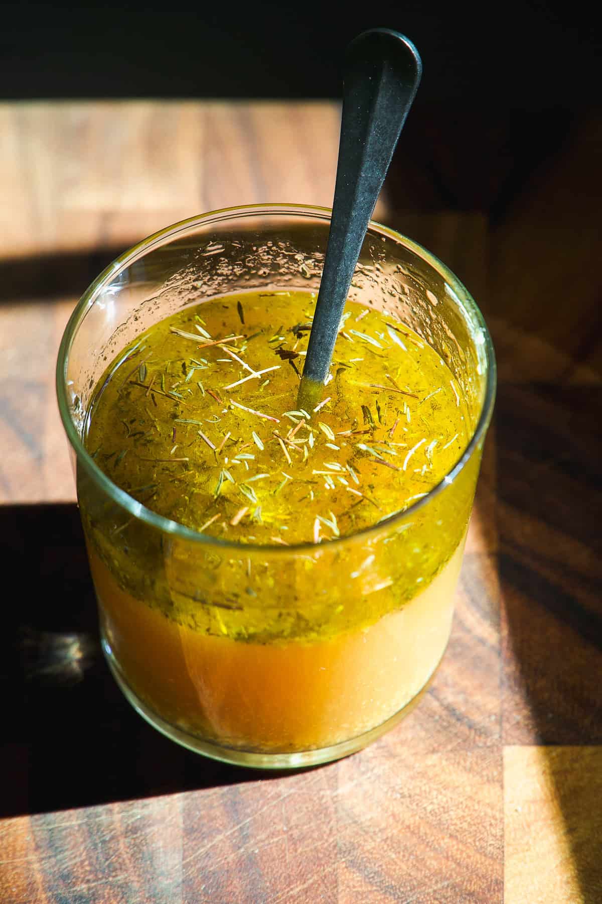 A clear glass filled with yellow vinaigrette salad dressing, topped with herbs, sits on a wooden surface with a spoon inside. Sunlight casts shadows and highlights the separation of oil and vinegar.
