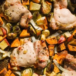 Baked chicken thighs on a sheet pan surrounded by roasted Brussels sprouts, sweet potatoes, onions, and apple chunks, all evenly browned and mixed together.