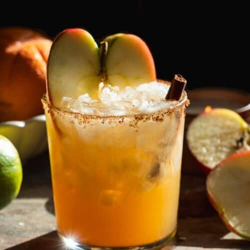 A glass of orange-colored cocktail with ice, garnished with two apple slices and a cinnamon stick, sits on a wooden surface with apples, a lime, and an orange in the background, bathed in warm light.