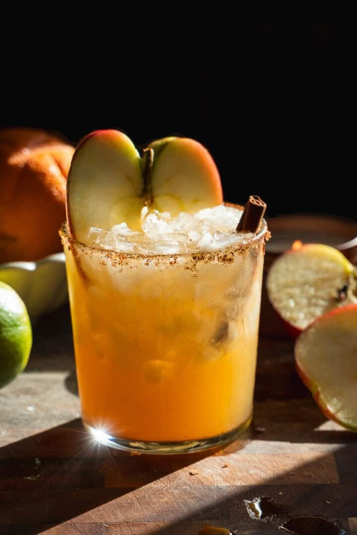 A glass of orange-colored cocktail with ice, garnished with two apple slices and a cinnamon stick, sits on a wooden surface with apples, a lime, and an orange in the background, bathed in warm light.