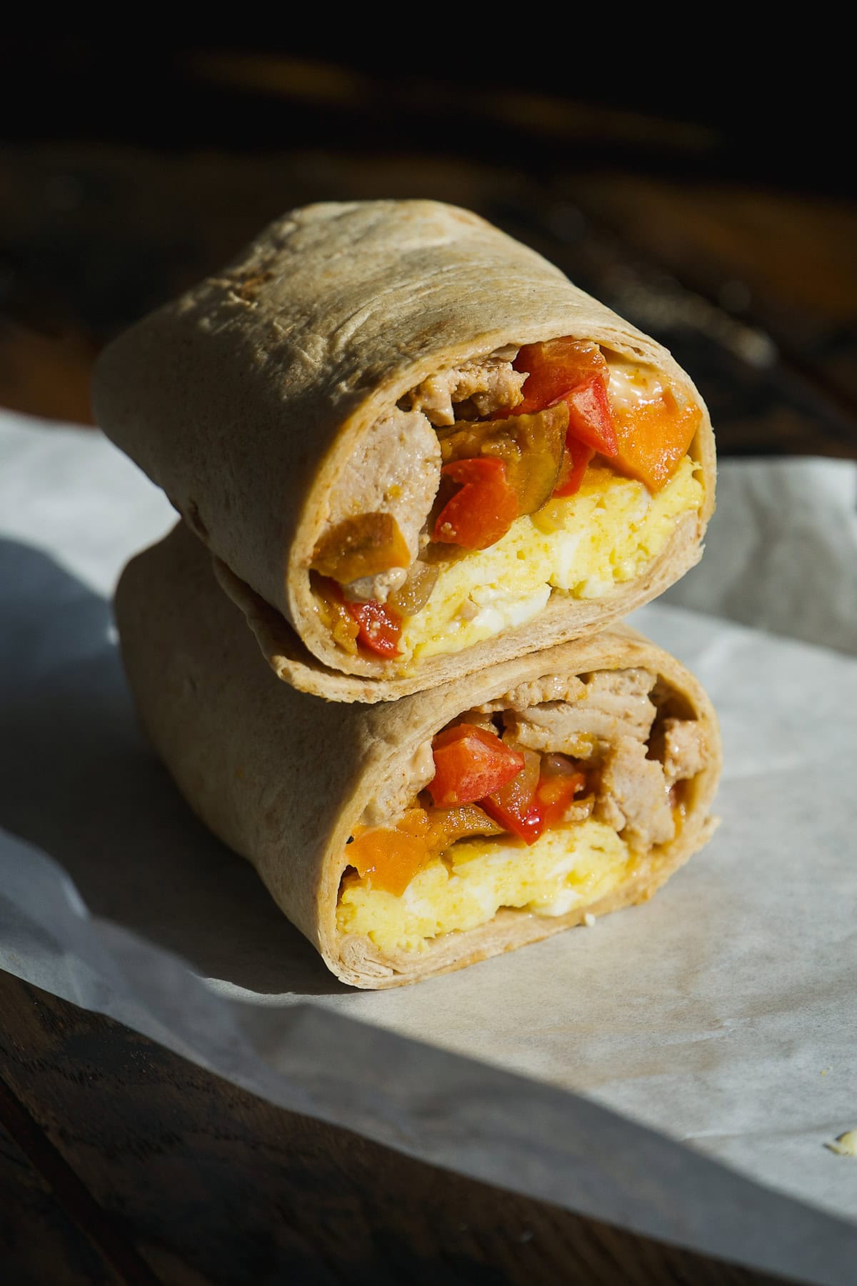 A breakfast burrito cut in half, stacked with visible scrambled eggs, sausage, and red bell peppers, rests on a sheet of parchment paper—perfect for make-ahead freezer breakfast burritos.