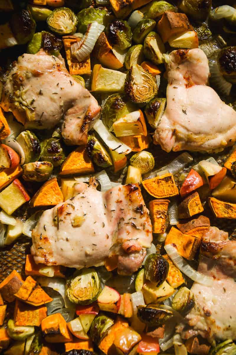 Roasted chicken thighs surrounded by chopped sweet potatoes, Brussels sprouts, and onions on a baking sheet, all cooked to a golden brown.