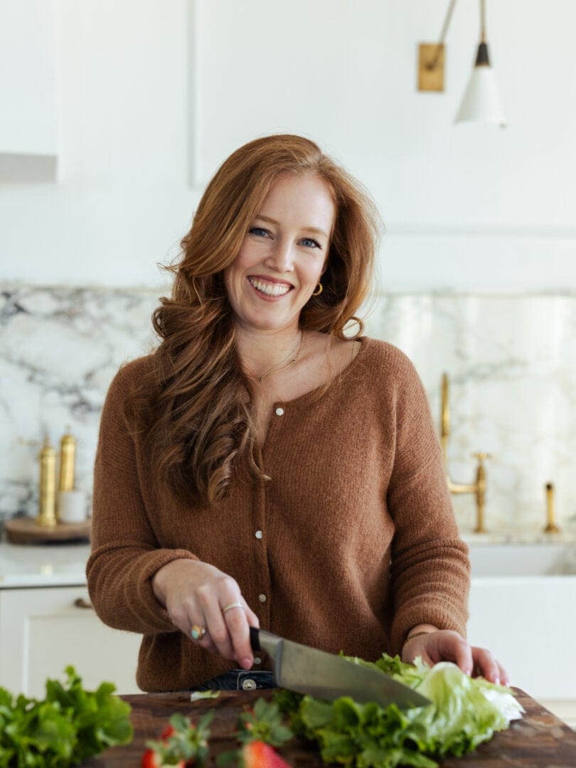 A woman with long red hair smiles while chopping lettuce on a wooden cutting board in a modern kitchen. Fresh greens and strawberries are on the countertop in front of her.