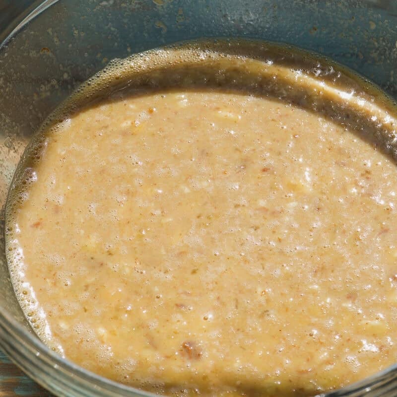 A glass bowl filled with a thick, light brown, and slightly foamy banana bread baked oats mixture rests on a wooden surface, capturing the moment this wholesome batter is being prepared.