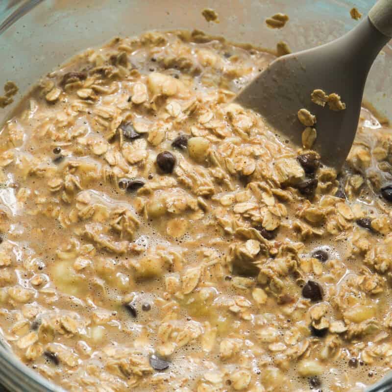 A glass bowl filled with a mixture for banana bread baked oats—oats, milk, chopped banana, and chocolate chips—is being stirred with a gray silicone spatula. The wet mixture looks perfect for baking or chilling.