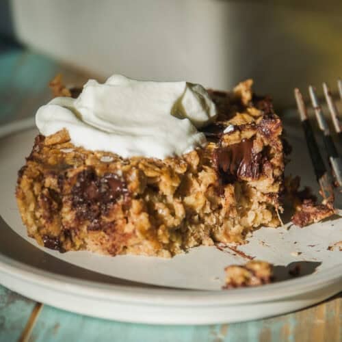 A close-up of banana bread baked oats with melted chocolate chunks, topped with a dollop of whipped cream, served on a plate with a fork beside it.