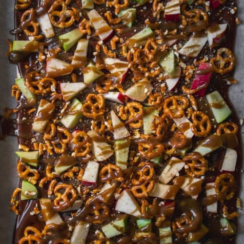 A sheet of chocolate bark topped with chopped apples, mini pretzels, and drizzles of caramel, displayed on parchment paper.